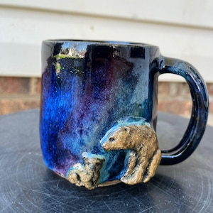 Puede incluir: Una taza de cerámica con un esmalte azul oscuro y púrpura. La taza presenta un diseño esculpido de oso y osezno en tonos marrones y dorados. El asa es de color azul oscuro a juego.
