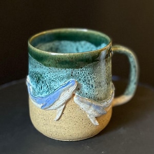 May include: Handmade ceramic mug with an ocean-themed design. The mug has a green rim, blue and green wave pattern, and two blue whale sculptures. The base is sandy beige, and the handle is green. The mug is approximately 10 cm tall.