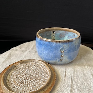 May include: A handmade ceramic garlic grater and bowl set. The bowl is a light blue with a brown rim and three small holes. The grater lid is tan with a textured surface. The set is on a neutral-coloured cloth.