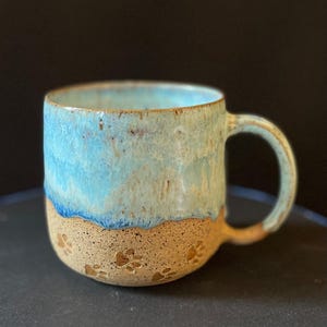 May include: A handmade ceramic mug with a light blue and tan glaze. The top half of the mug is light blue, transitioning to tan at the bottom. Paw prints are imprinted on the tan base. The mug has a curved handle.