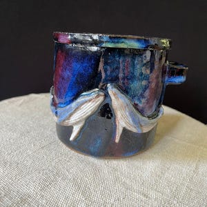 May include: A handmade ceramic mug with a dark blue, purple, and black glaze. Two white and grey whale figures are sculpted on the mug's exterior. The mug has a small handle and a textured rim.