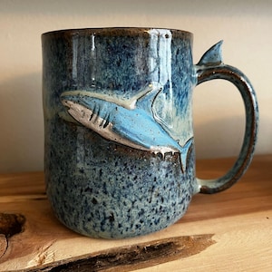 Puede incluir: Una taza de cerámica con un esmalte azul y marrón, con un relieve en relieve de un tiburón. El tiburón es azul claro con detalles blancos. El asa tiene un detalle en forma de aleta. La taza está sobre una superficie de madera.