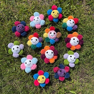May include: A collection of colorful, crocheted flower-shaped plushies with smiling faces. Each flower features a different color combination, including blue, pink, yellow, red, and purple. The plushies are arranged on a bed of green grass.