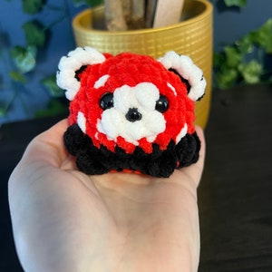 May include: A small crocheted red panda plush toy with black and white details. The toy has large black eyes and a white nose.