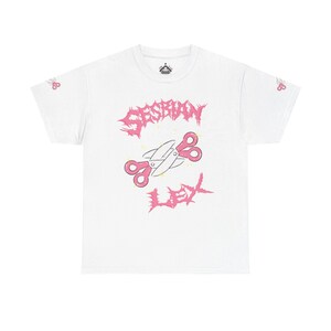 May include: A white short-sleeve t-shirt featuring a pink graphic of crossed scissors in the center. The text "SESBIAN" appears above the scissors and "LEX" below in a jagged pink font. Small pink scissors are printed on each sleeve.