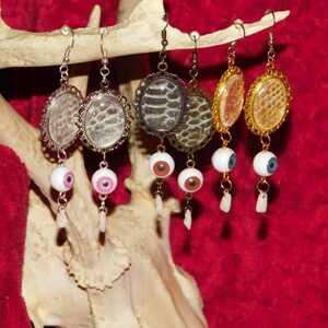 May include: Three pairs of earrings with a snakeskin pattern and dangling eyeballs. The earrings are hanging from a deer antler.