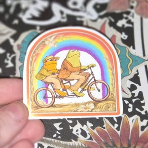 May include: A die-cut sticker featuring an illustration of two anthropomorphic frogs riding a tandem bicycle. The frogs are wearing tan jackets and hats, positioned under a vibrant, multi-colored rainbow arch. The background shows a simple landscape.