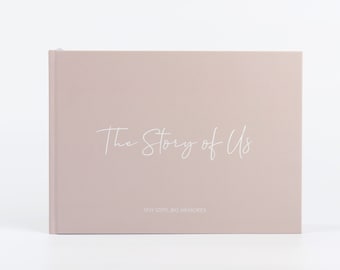 Może przedstawiać: Różowy, twardo oprawiony dziennik z napisem "The Story of Us" w białym kolorze i tekstem "Tiny Steps, Big Memories" w mniejszym białym kolorze poniżej.