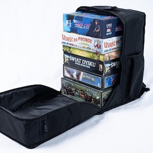 Peut inclure: Un sac à dos noir, ouvert en haut, rempli de plusieurs boîtes de jeux de société. Les boîtes sont empilées et présentent des graphiques et du texte colorés. Le sac à dos a une poignée et une poche latérale. Le compartiment ouvert est au premier plan.