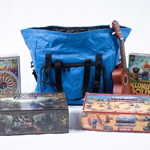 Könnte beinhalten: Eine blaue Tragetasche mit schwarzen Riemen und eine braune Ukulele neben Brettspielen. Zu den Brettspielen gehören "Sagrada", "A Song of Ice & Fire", "Masters of the Universe" und "Klonowa Dolina".