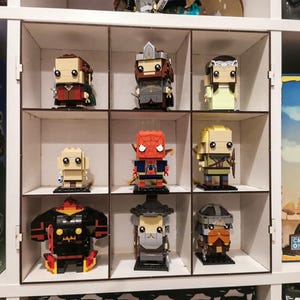 May include: A white shelf unit with nine square compartments, each displaying a different Lego minifigure. The minifigures include characters from popular franchises like Lord of the Rings, Marvel, and Star Wars.