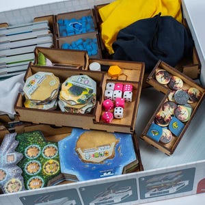 May include: A board game organiser with various components, including cards, tokens, dice, and wooden dividers. The game pieces are arranged in a white box, with blue, yellow, and pink accents.