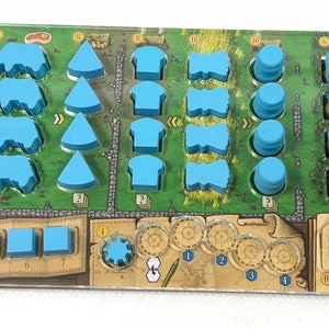 以下が含まれることがあります： 透明なプラスチック製のゲームボードに、さまざまな形をした青い木製のパーツが置かれています。ボードは緑と茶色の背景に地図のデザインが施されています。パーツは行と列に配置されています。ボードには数字と記号もいくつかあります。