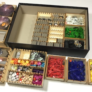 Puede incluir: Un conjunto de organizadores de madera para el almacenamiento de juegos de mesa. Los organizadores están hechos de madera marrón claro y tienen varios compartimentos para guardar diferentes piezas de juego. Los organizadores están llenos de una variedad de piezas de juego, incluyendo dados, fichas y cartas.