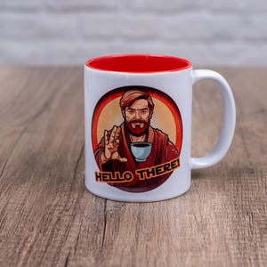 May include: White ceramic mug with a red rim. The mug features a cartoon image of a man with a beard and brown hair, holding a cup and saying "Hello There!"