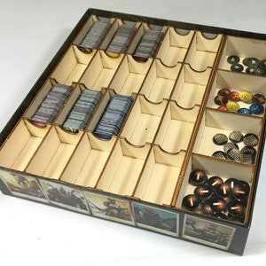 May include: A wooden organizer with multiple compartments for storing game pieces and cards. The organizer has a brown and black exterior with images of characters from a game. The organizer is filled with various game pieces, including tokens, cards, and dice.