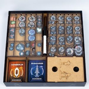 Puede incluir: Un organizador de juegos de mesa negro con múltiples compartimentos llenos de piezas de juego, cartas y un marcador. Las cartas tienen el texto "CERBERUS" y "ZAGROZENIE". Las piezas del juego son de varios colores y formas.