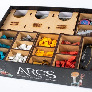 Op de afbeelding: Een zwarte speldoos met houten verdelers, gevuld met kleurrijke spelstukken. De doos is gelabeld "ARCS: Conflict & Collapse in the Reach". Compartimenten bevatten blauwe, witte, gele en rode spelcomponenten, samen met kaarten en tokens.