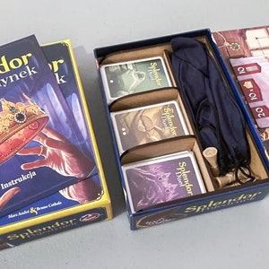 Może przedstawiać: Niebieskie pudełko z grą w środku. Na pudełku widnieje napis "Splendor Pojednynek". Gra zawiera niebieski worek, drewniane żetony i karty z napisem "Splendor Duel".