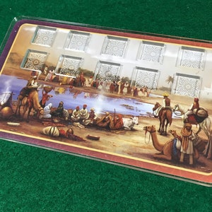Puede incluir: Un tablero de juego rectangular con una ilustración colorida de una escena del desierto con camellos, personas y un cuerpo de agua. El tablero tiene 10 cuadrados de plástico transparente con patrones decorativos.