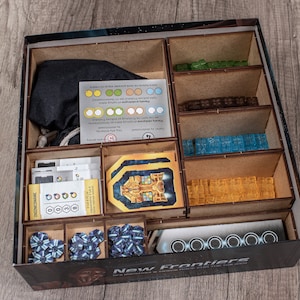 May include: A wooden box organizer for the board game "New Frontiers". The box has compartments for storing the game's components, including cards, tiles, and tokens. The box is made of wood and has a black and white design.