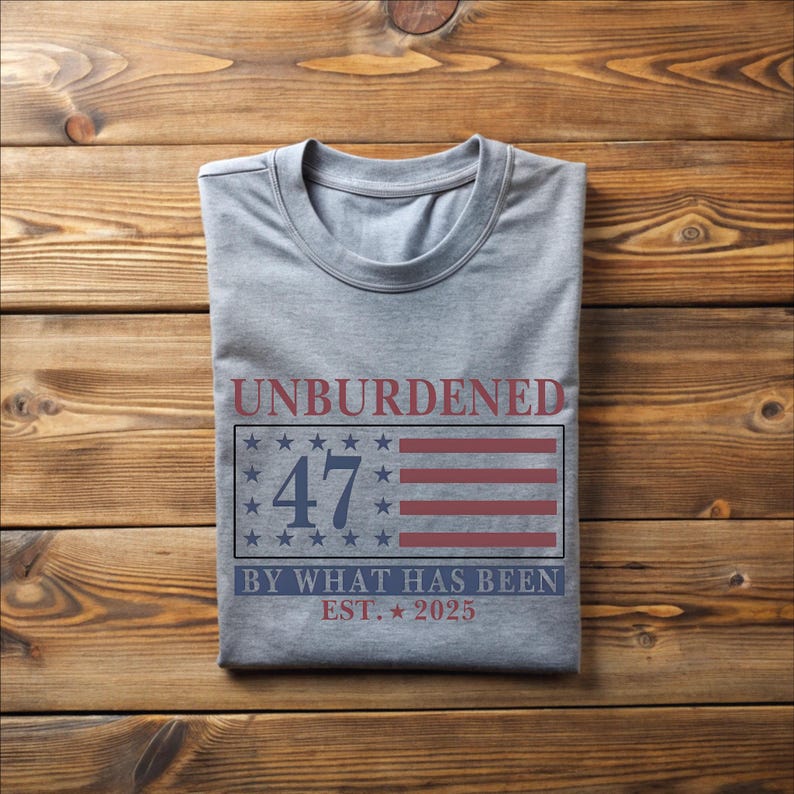 Puede incluir: Una camiseta gris con un dise&ntilde;o de bandera estadounidense roja, blanca y azul. El texto "Unburdened 47 By What Has Been Est. 2025" est&aacute; impreso en la camiseta.
