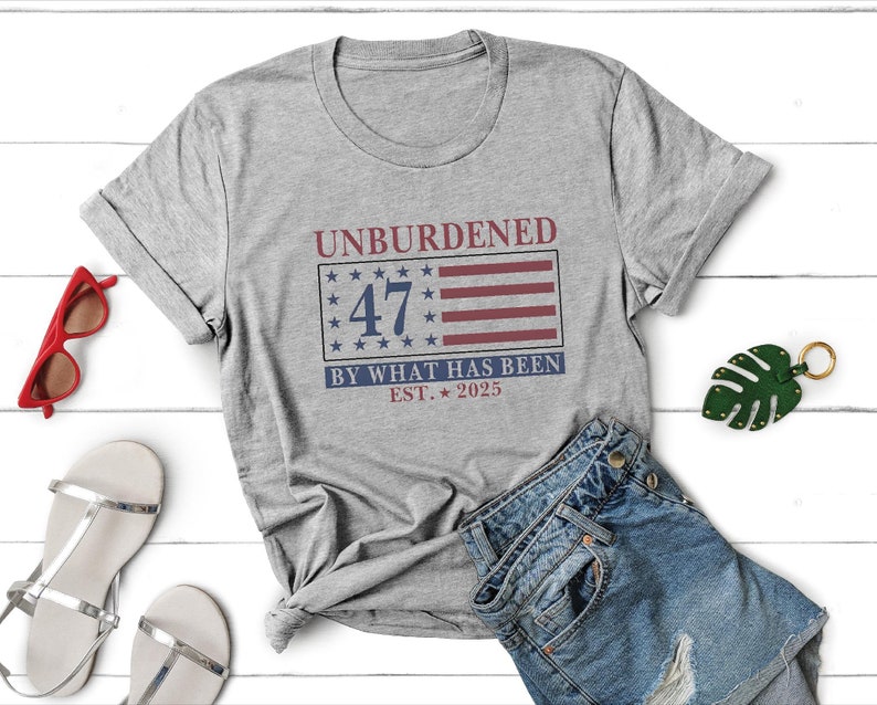 Puede incluir: Camiseta gris con un dise&ntilde;o de bandera americana roja, blanca y azul. El texto "Unburdened 47 By What Has Been Est. 2025" est&aacute; impreso en la camiseta.