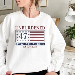 Puede incluir: Sudadera blanca con un dise&ntilde;o de bandera americana roja, blanca y azul. El texto "UNBURDENED 47 BY WHAT HAS BEEN EST. 2025" est&aacute; impreso en la sudadera.