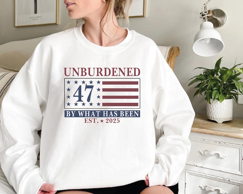 Puede incluir: Sudadera blanca con un dise&ntilde;o de bandera americana roja, blanca y azul. El texto "UNBURDENED 47 BY WHAT HAS BEEN EST. 2025" est&aacute; impreso en la sudadera.