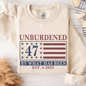 Puede incluir: Una sudadera beige con el texto "Unburdened 47 By What Has Been Est. 2025" impreso en la parte delantera. El texto est&aacute; en azul marino con un gr&aacute;fico de bandera estadounidense roja, blanca y azul detr&aacute;s del n&uacute;mero 47.