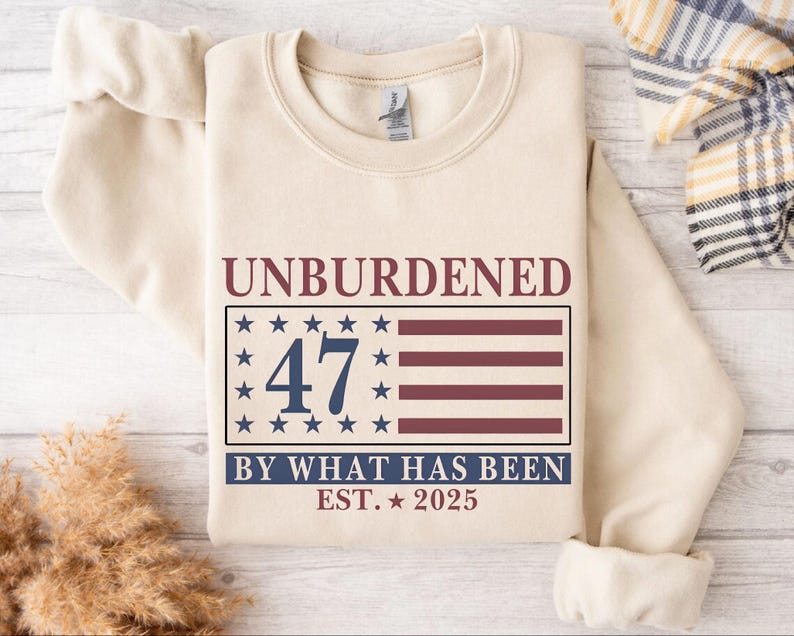Puede incluir: Una sudadera beige con el texto "Unburdened 47 By What Has Been Est. 2025" impreso en la parte delantera. El texto est&aacute; en azul marino con un gr&aacute;fico de bandera estadounidense roja, blanca y azul detr&aacute;s del n&uacute;mero 47.