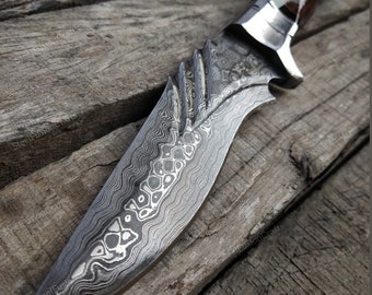 May include: A close-up of a Damascus steel knife with a wooden handle. The blade has a wavy pattern and a sharp point. The handle is brown and has a smooth finish.