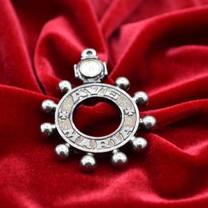 May include: A silver-toned pendant with the words "AVE MARIA" inscribed around a central circle. The pendant has a circular design with small, spherical accents and is set against a red velvet background.