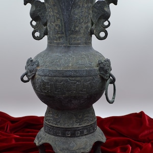 Puede incluir: Un jarrón de bronce gris oscuro con tallas intrincadas y asas en forma de pájaro. El jarrón tiene un cuerpo redondo y una base de tres patas.