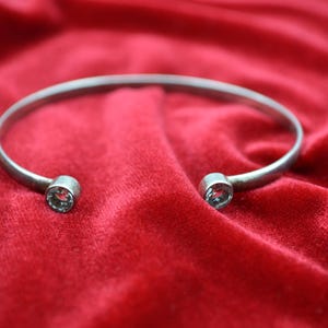 Puede incluir: Una pulsera de puño abierta plateada con una gema en cada extremo. La pulsera se exhibe sobre una superficie de terciopelo rojo. Las gemas parecen claras, añadiendo un toque de brillo a la joya.
