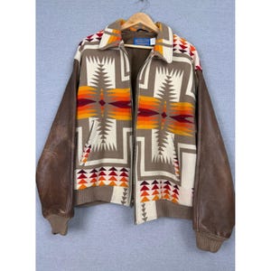 May include: A zip-up jacket featuring brown leather sleeves and a tan wool body with a geometric pattern in orange, red, and cream. The jacket includes a collar, ribbed cuffs, and a Pendleton brand label on the inside neck area.