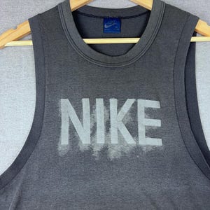 Puede incluir: Una camiseta sin mangas gris con la palabra "NIKE" en blanco en la parte delantera. La camiseta sin mangas está colgada de una percha de madera. Una pequeña etiqueta azul con el logotipo de Nike es visible cerca del cuello.