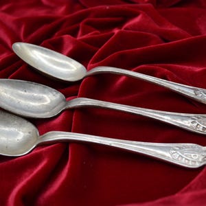 May include: Three silver spoons with decorative handles are arranged on a deep red velvet cloth. The spoons have a traditional design, with a rounded bowl and an ornate pattern at the end of the handle.