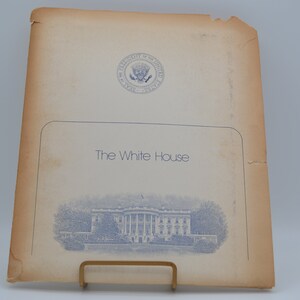 Op de afbeelding: Een witte map met het presidentiële zegel van de Verenigde Staten en de tekst "The White House" in het blauw gedrukt. Een blauwe en witte illustratie van het Witte Huis is afgedrukt op de voorkant van de map.
