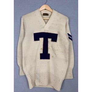 May include: A cream-colored V-neck sweater featuring a large, dark navy blue letter T appliqued on the chest. The long-sleeved knit garment includes ribbed cuffs and a ribbed hem, with two dark navy blue horizontal stripes on the left sleeve.