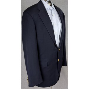 Peut inclure: Un blazer bleu marine doté d'un revers cranté, de deux boutons en métal doré sur le devant et de trois boutons dorés plus petits sur les poignets. Le blazer est porté sur une chemise bleu clair à col pointu et boutonnée sur le devant.