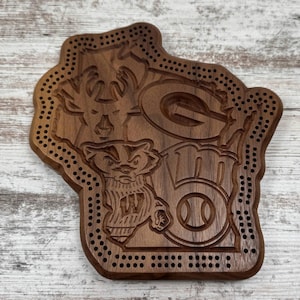 May include: A wooden cribbage board in the shape of Wisconsin, featuring carved images of a deer, a badger, a baseball, and the Green Bay Packers logo. The board has numerous holes for pegs and is made of dark brown wood.