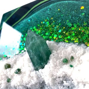 Op de afbeelding: Een groen kristal op een witte, glinsterende achtergrond. Het kristal is omgeven door kleinere groene kristallen.