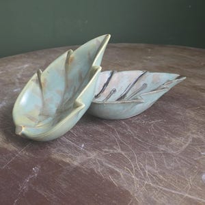 May include: Two ceramic leaf-shaped bowls. The bowls are a pale green with darker green and brown accents. One bowl is in the foreground, and the other is behind it. The bowls are on a brown wooden surface.