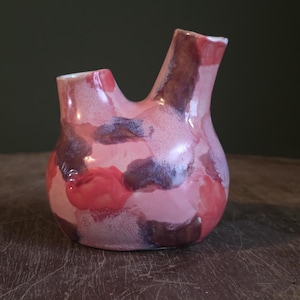 Puede incluir: Un jarrón de cerámica hecho a mano con una forma única y un acabado brillante. El jarrón está pintado con una mezcla de tonos rosas, rojos y morados, creando un efecto acuarela. Tiene dos aberturas, una en la parte superior y otra en el lateral.