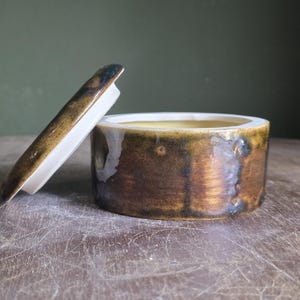 May include: A small, round ceramic container with a lid. The container is a mix of brown and gold tones, with a white interior. The lid is partially open, revealing the inside. The piece is on a wooden surface.