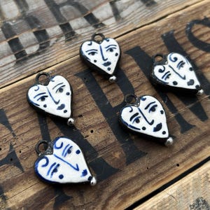 May include: Five heart-shaped ceramic charms with a face design. The charms are white with blue facial features and black outlines. Each charm has a small silver-colored bead at the bottom and a loop at the top for hanging. They rest on a wooden surface.