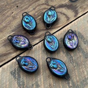 May include: Seven iridescent oval pendants with a black border and a tulip design. Each pendant has a loop at the top for hanging. The tulip design is in shades of blue, purple, and green, with a glossy finish. The pendants are on a wooden surface.