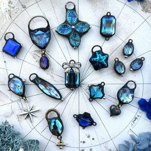 May include: A collection of blue and iridescent glass pendants and charms. The selection features diverse shapes such as squares, teardrops, and stars, with metal frames and loops for jewellery making or crafting. The items are displayed on a map-like background.