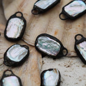 May include: Close-up of several irregularly shaped pendants with a pearlescent center and a dark, possibly oxidized, metal frame. Each pendant has a small loop at the top. The pendants are arranged on a textured, off-white surface.